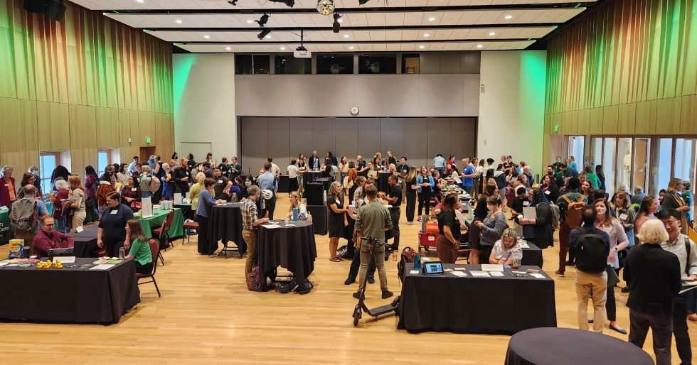 A wide view of a large indoor resource fair where many people stand and talk around tables with informational displays in a modern hall with wood floors and tall walls lit with green lighting.